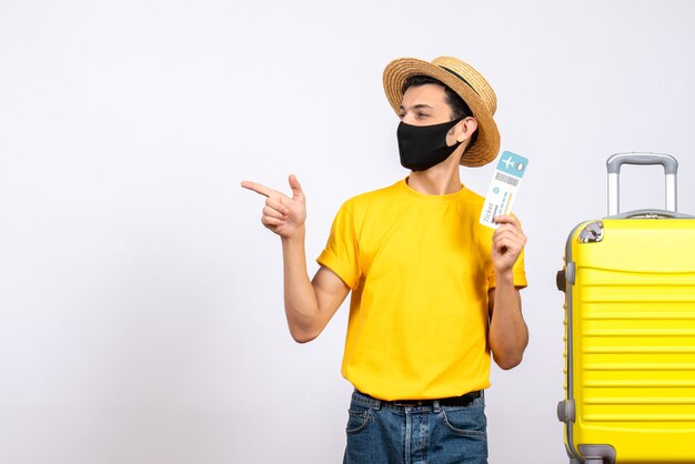 Vue de face jeune homme avec chapeau de paille et masque debout près de valise jaune tenant un billet de voyage pointant vers la gauche