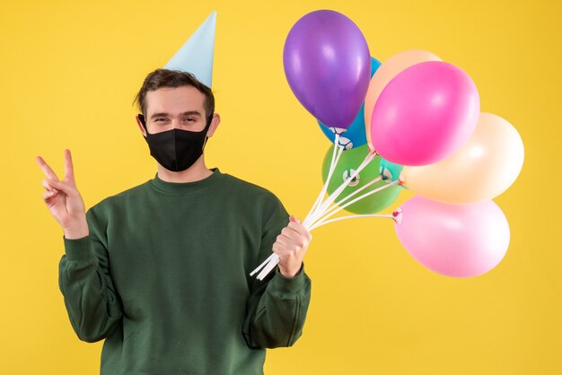 Vue de face jeune homme avec chapeau de parti faisant signe de victoire debout sur fond jaune