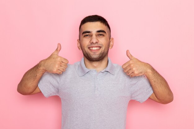 Vue de face jeune homme en chemise grise posant et souriant montrant des gestes horribles sur rose