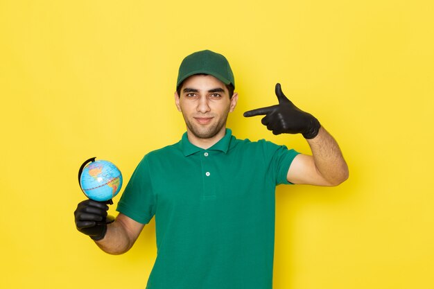 Vue de face jeune homme en chemise verte casquette verte tenant petit globe avec des gants noirs sur jaune