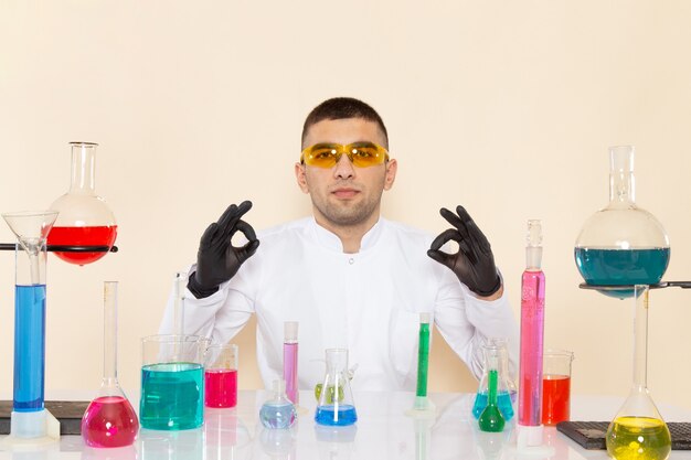 Vue de face jeune homme chimiste en costume spécial blanc assis en face de la table avec des solutions sur l'expérience scientifique de chimie de laboratoire mur crème