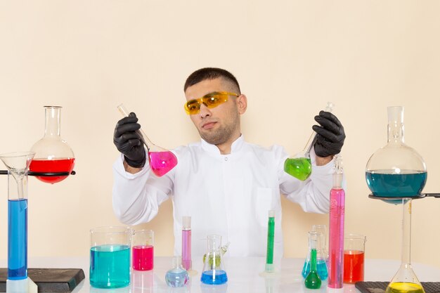 Vue de face jeune homme chimiste en costume spécial blanc assis en face de la table avec des solutions tenant sur une expérience scientifique de chimie de laboratoire de mur de crème