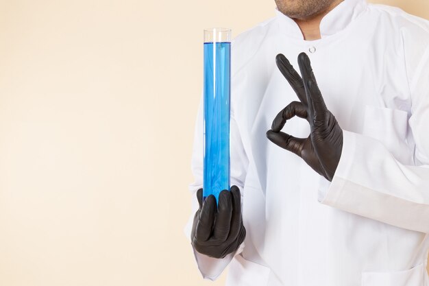 Vue de face jeune homme chimiste en costume spécial blanc tenant un petit flacon avec une solution bleue sur mur crème laboratoire chimie expérience scientifique