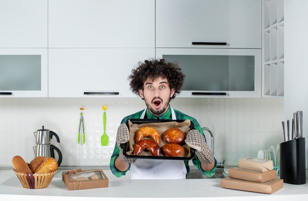 Vue de face d'un jeune homme choqué portant un support montrant une pâtisserie fraîchement préparée dans la cuisine blanche