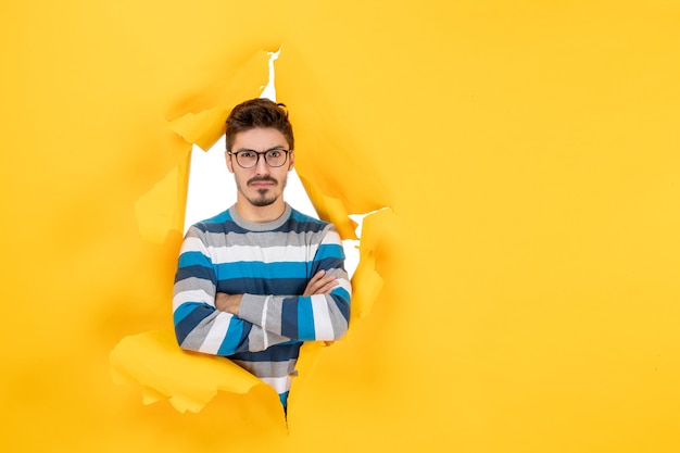 Vue de face jeune homme confus regardant à travers un mur jaune en papier déchiré