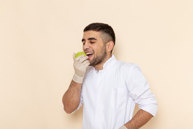 Vue de face jeune homme en costume blanc portant des gants mordant la pomme sur beige