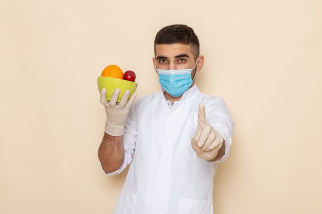 Vue de face jeune homme en costume blanc portant un masque et des gants tenant la plaque avec des fruits sur beige