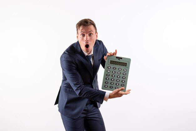 Vue de face jeune homme en costume classique élégant tenant une énorme calculatrice sur un bureau blanc