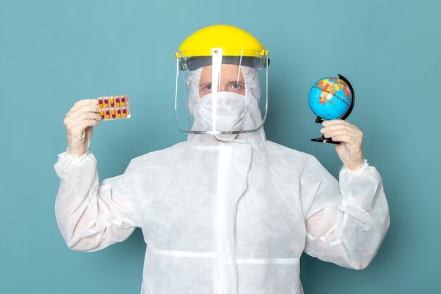 Une vue de face jeune homme en costume spécial blanc et casque spécial jaune tenant petit globe et pilules sur le mur bleu homme costume danger couleur d'équipement spécial