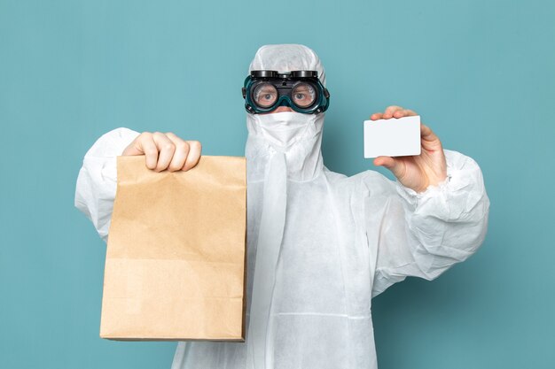 Une vue de face jeune homme en costume spécial blanc et tenant une carte blanche et un paquet sur le mur bleu homme costume couleur équipement spécial danger