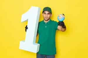 Photo gratuite vue de face jeune homme courrier en chemise verte casquette verte gants noirs tenant petit globe et numéro un sur jaune