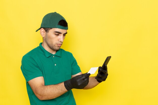 Vue de face jeune homme courrier en chemise verte casquette verte tenant un smartphone carte blanche sur jaune