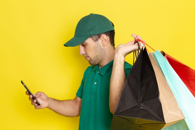 Vue de face, jeune, homme, courrier, dans, chemise verte, chapeau vert, tenue, achats, paquets, et, utilisation, a, téléphone, sur, jaune