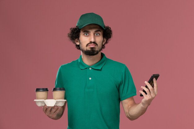 Vue de face jeune homme courrier en uniforme vert et cape tenant des tasses de café de livraison et son téléphone en pensant sur fond rose service travail de livraison uniforme