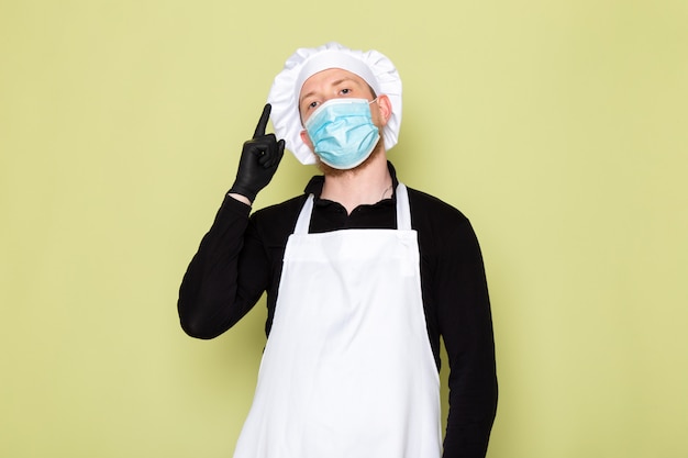 Une vue de face jeune homme cuisinier en chemise noire avec cape blanche tête blanche en gants noirs masque de protection bleu