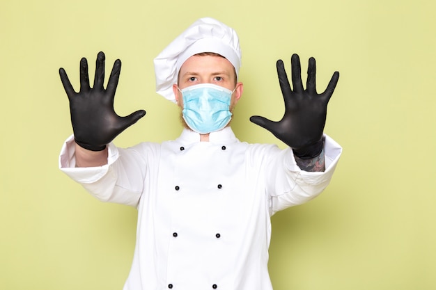 Photo gratuite une vue de face jeune homme cuisinier en costume de cuisinier blanc casquette blanche dans des gants noirs masque de protection bleu montrant ses mains