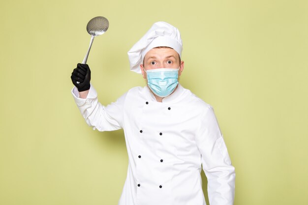 Une vue de face jeune homme cuisinier en costume de cuisinier blanc casquette blanche dans des gants noirs masque de protection bleu tenant une grosse cuillère métallique menaçant