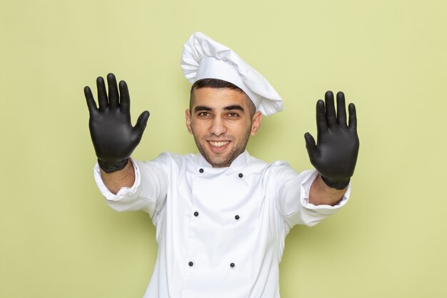 Vue de face jeune homme cuisinier en costume de cuisinier blanc avec des gants noirs montrant ses mains sur le vert