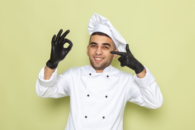 Vue de face jeune homme cuisinier en costume de cuisinier blanc portant des gants en cuir noir avec sourire sur vert