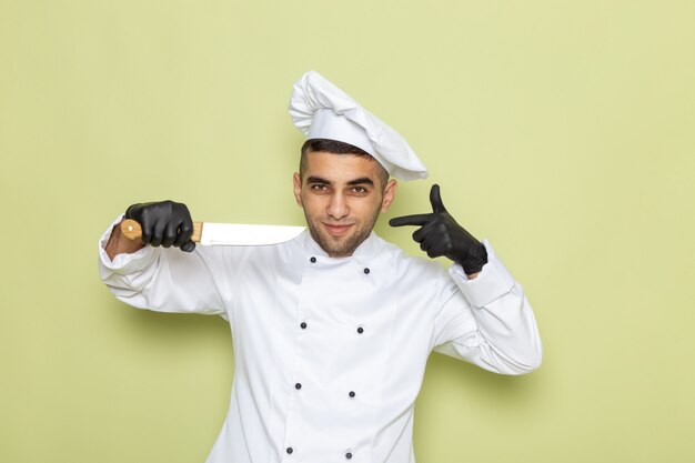 Vue de face jeune homme cuisinier en costume de cuisinier blanc portant des gants noirs et tenant un couteau sur vert