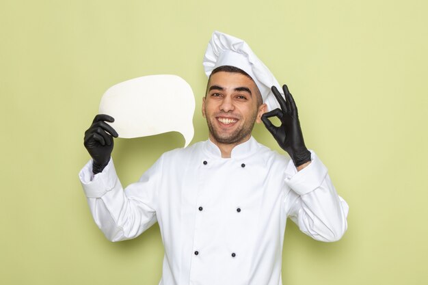 Vue de face jeune homme cuisinier en costume de cuisinier blanc portant des gants noirs tenant panneau blanc sur vert