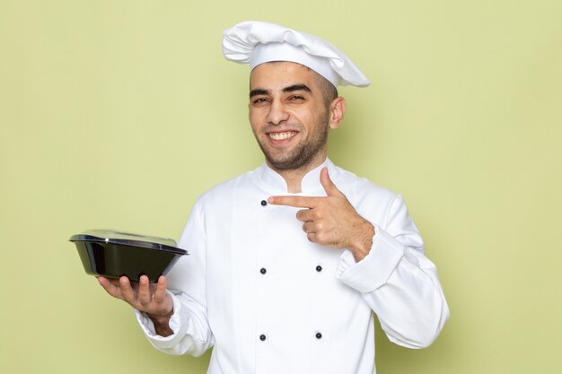 Vue de face jeune homme cuisinier en costume de cuisinier blanc tenant un bol alimentaire noir et souriant sur vert