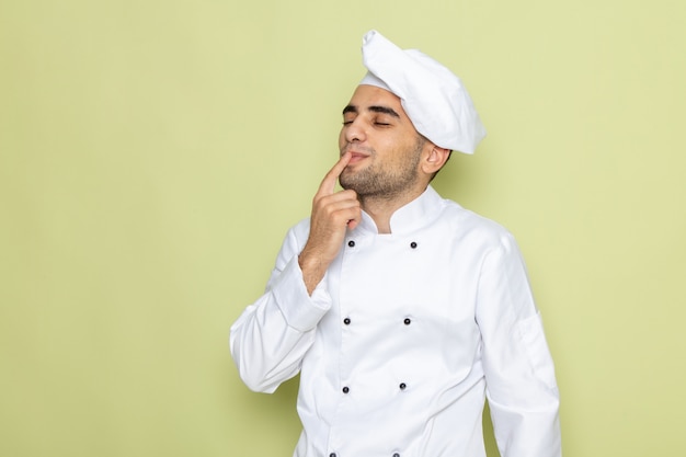 Vue de face jeune homme cuisinier en costume de cuisinier blanc avec les yeux fermés montrant un signe savoureux sur vert