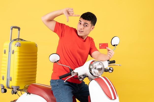 Vue de face jeune homme sur cyclomoteur avec valise jaune holding card