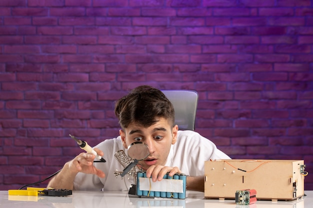 Vue de face jeune homme derrière le bureau essayant de réparer la petite disposition de la maison sur le mur violet