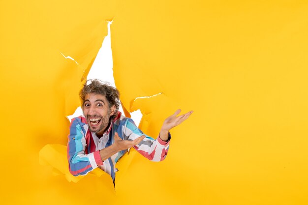 Vue de face d'un jeune homme émotif et fou posant pour la caméra à travers un trou déchiré dans du papier jaune