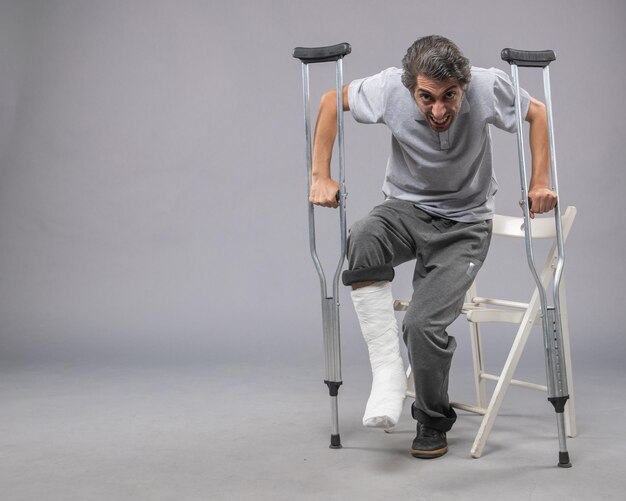 Vue de face jeune homme essayant de se lever avec un pied cassé à l'aide de béquilles sur des jambes de mur gris torsion accident douleur pied cassé