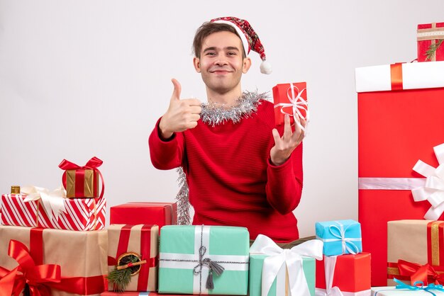 Vue de face jeune homme faisant signe de pouce vers le haut assis autour de cadeaux de Noël