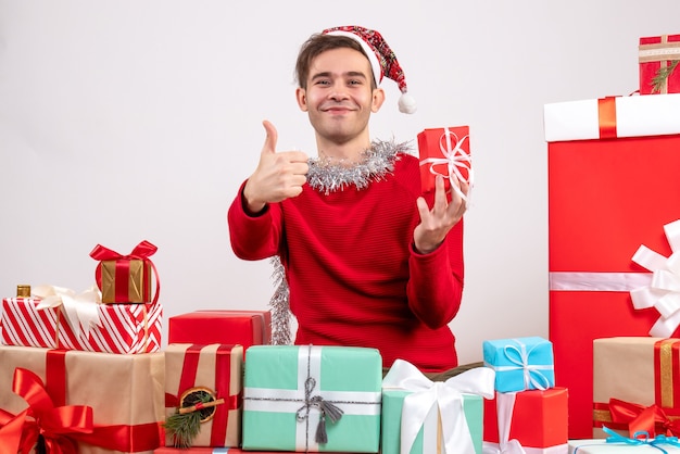 Vue de face jeune homme faisant signe de pouce vers le haut assis autour de cadeaux de Noël