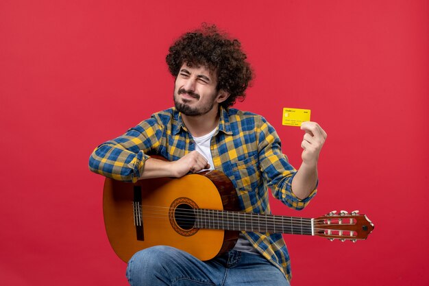 Vue de face jeune homme avec guitare tenant une carte bancaire jaune sur un mur rouge applaudissements de performance concert live