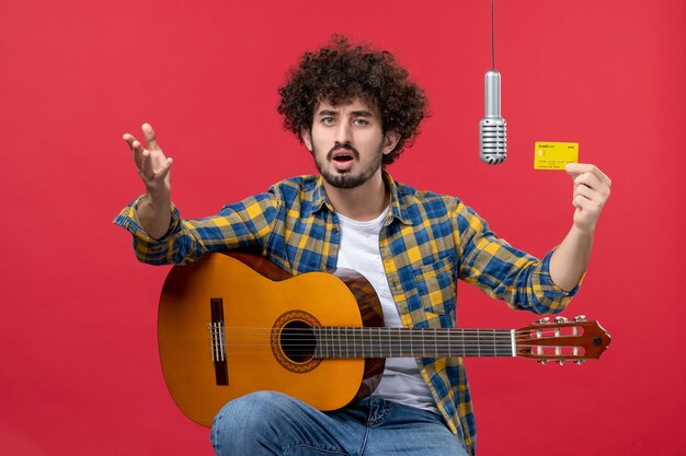 Vue de face jeune homme avec guitare tenant une carte bancaire sur le mur rouge live couleur performance musicien bande argent