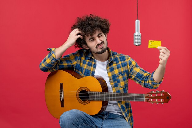 Vue de face jeune homme avec guitare tenant une carte bancaire sur le mur rouge live couleur performance musicien concert argent