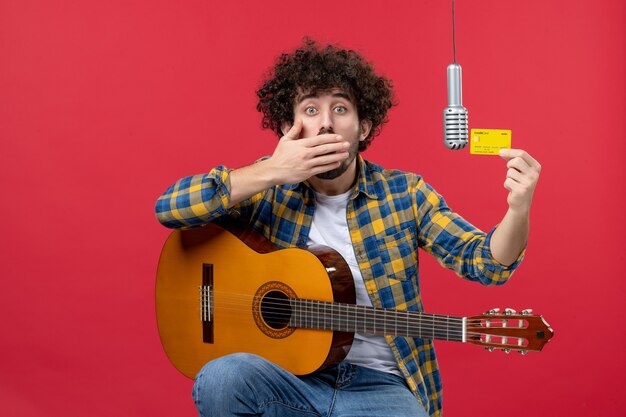 Vue de face jeune homme avec guitare tenant une carte bancaire sur le mur rouge live performance musicien concert argent couleur