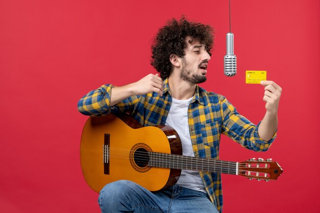 Vue de face jeune homme avec guitare tenant une carte bancaire sur le mur rouge live performance musicien concert band argent couleur