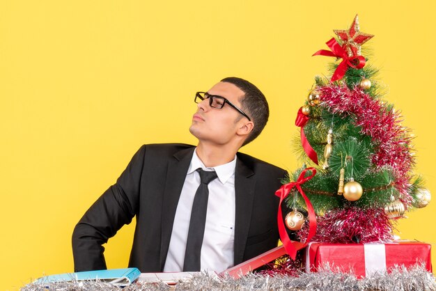 Vue de face jeune homme avec des lunettes assis à la table en regardant à gauche arbre de Noël et cadeaux