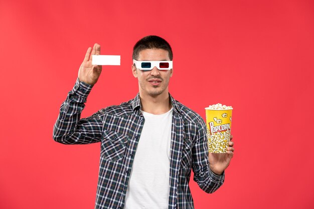 Vue de face jeune homme en -d lunettes de soleil tenant le paquet de pop-corn et billet sur mur rouge clair cinéma films cinéma film mâle