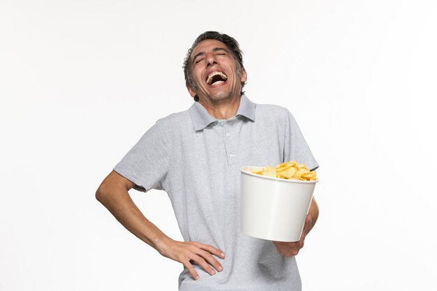 Vue de face jeune homme mangeant des chips de pomme de terre en riant sur une surface blanche