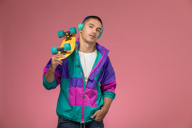 Photo gratuite vue de face jeune homme en manteau coloré, écouter de la musique tenant une planche à roulettes sur un bureau rose