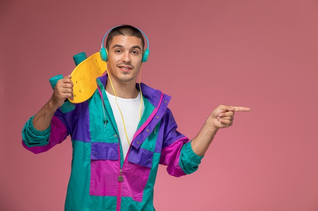 Vue de face jeune homme en manteau coloré, écouter de la musique tenant une planche à roulettes sur un bureau rose