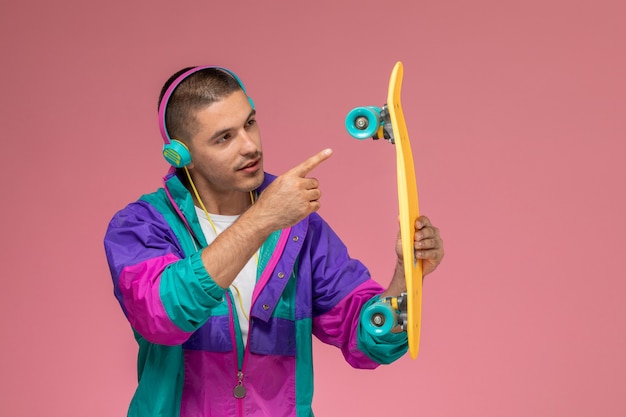 Vue de face jeune homme en manteau coloré, écouter de la musique et tenir la planche à roulettes sur un bureau rose clair