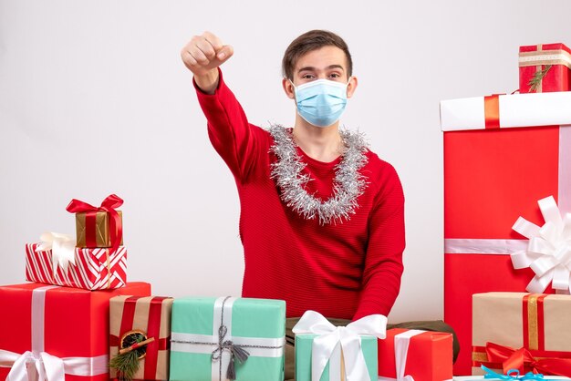 Vue de face jeune homme avec masque assis autour de cadeaux de Noël blanc