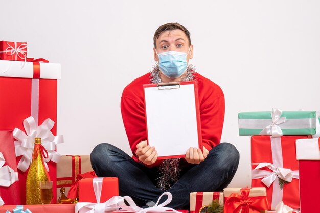 Photo gratuite vue de face d'un jeune homme en masque assis autour de cadeaux de noël sur un mur blanc