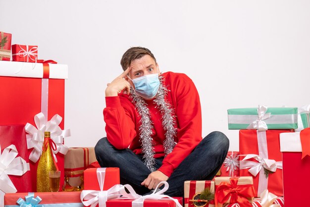 Vue de face d'un jeune homme en masque assis autour de cadeaux de Noël sur un mur blanc