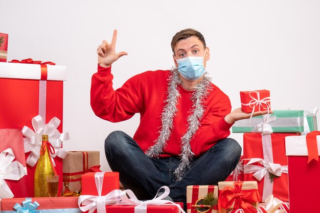 Vue de face d'un jeune homme en masque assis autour de cadeaux de Noël sur un mur blanc