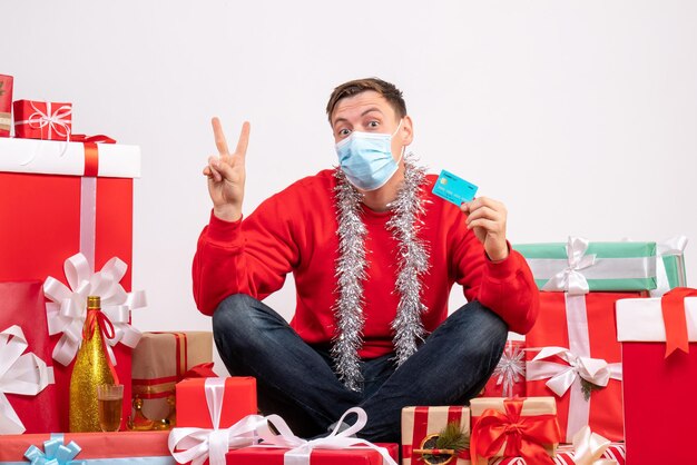 Vue de face d'un jeune homme en masque assis autour de cadeaux de Noël sur un mur blanc