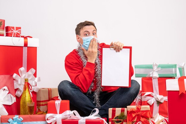 Vue de face d'un jeune homme en masque assis autour de cadeaux de Noël avec une note sur un mur blanc